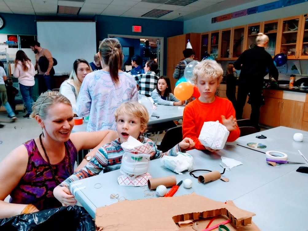 Many people of all ages inside a classroom, building vehicles for an Egg Drop Challenge