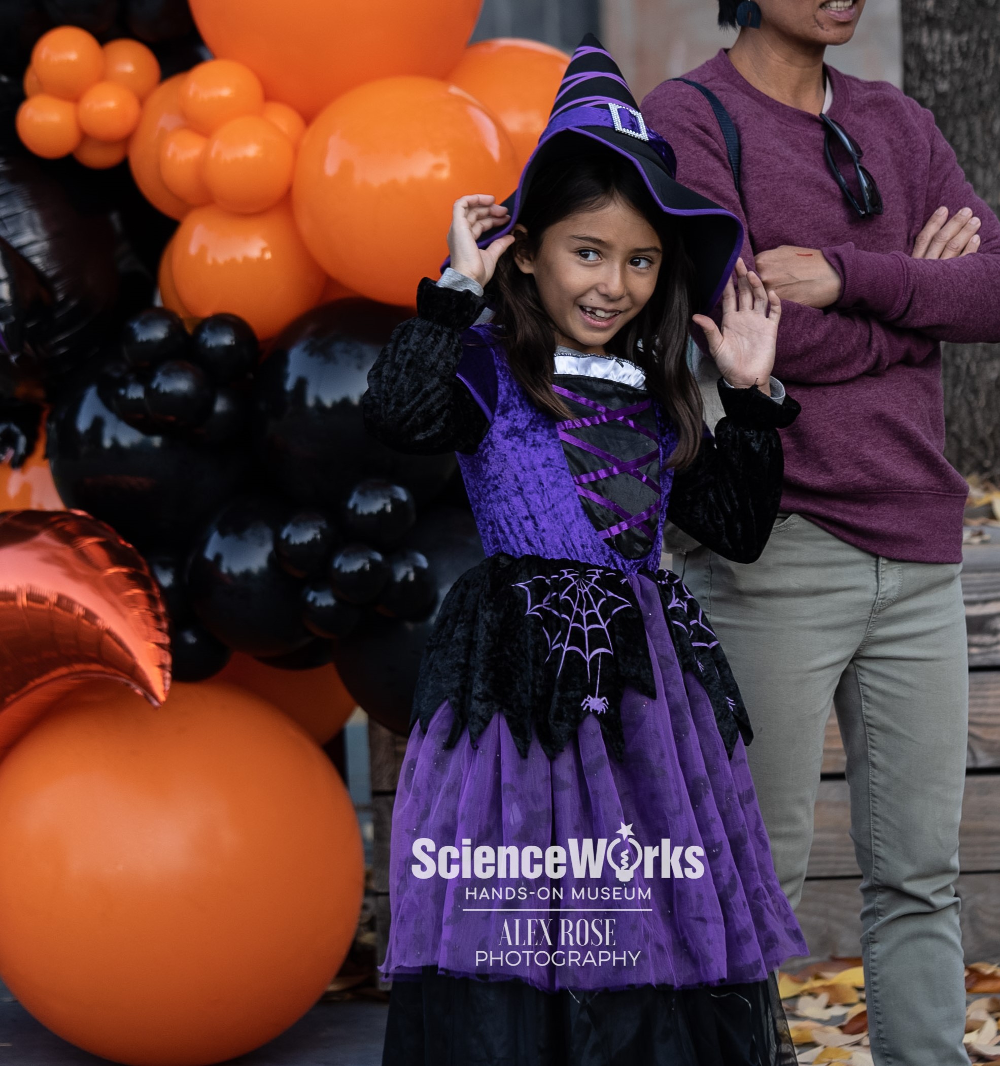 Young smiling girl dressed as a purple witch with black and orange balloons around her